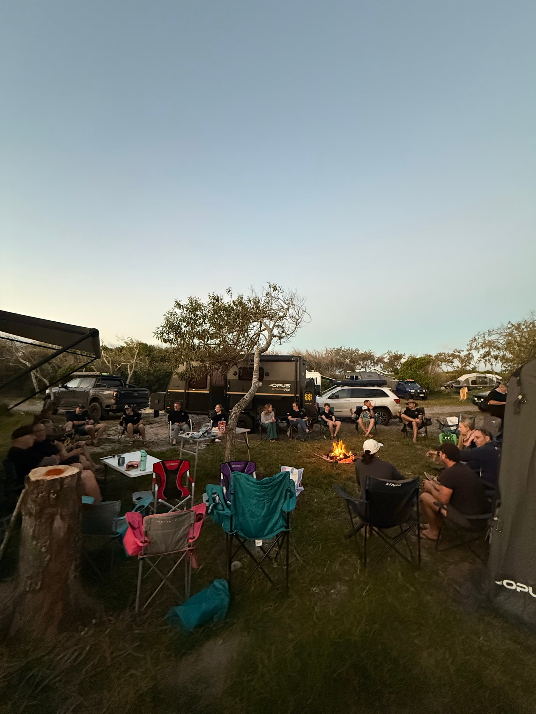 OPUS owners gathered around a campfire at dusk, campers parked behind